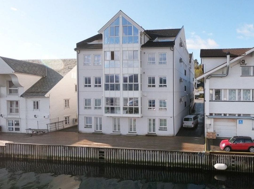 A multi-story building is displayed with large glass windows, allowing natural light to fill the interior. The exterior is clad in white, and a deck with wooden planks can be seen along the waterfront. Nearby structures feature traditional architectural elements.