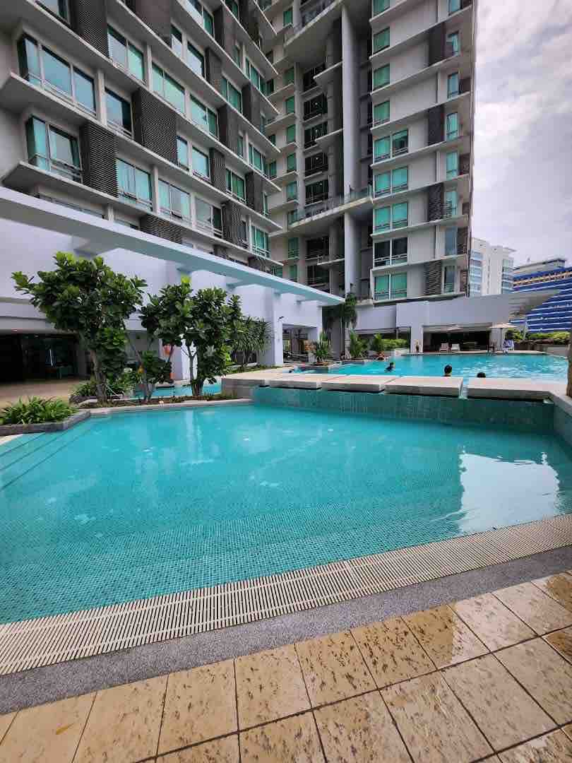 A serene swimming pool is presented, surrounded by lush greenery and modern architecture. The clear water reflects the sky, while lounge areas are visible in the background. Sun loungers are arranged near the pool, providing a relaxing space for guests.