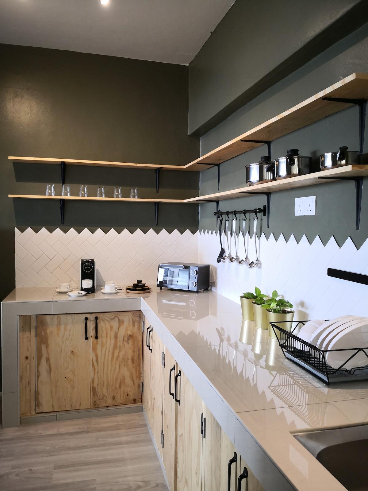 The kitchen features a modern design with light wood cabinetry and open shelving. A coffee maker and microwave are neatly arranged on the countertop, along with decorative plants. Glassware and kitchen utensils are displayed above, complementing the minimalist aesthetic.
