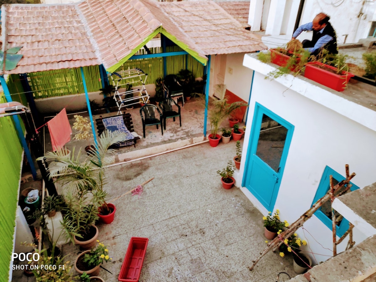 An open courtyard is visible, surrounded by vibrant potted plants and seating areas. A semi-covered porch provides shade, with a clothes drying rack positioned nearby. Brightly painted walls enhance the lively atmosphere, while a gardener can be seen tending to the terrace plants.