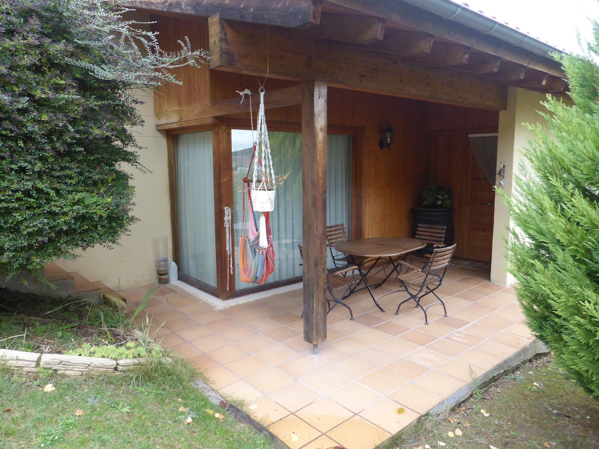 A sheltered patio area features a round wooden table accompanied by several black metal chairs. The smooth tiled floor leads to large glass doors, which provide access to the interior. Lush greenery and shrubs frame the entrance, enhancing the natural surroundings.