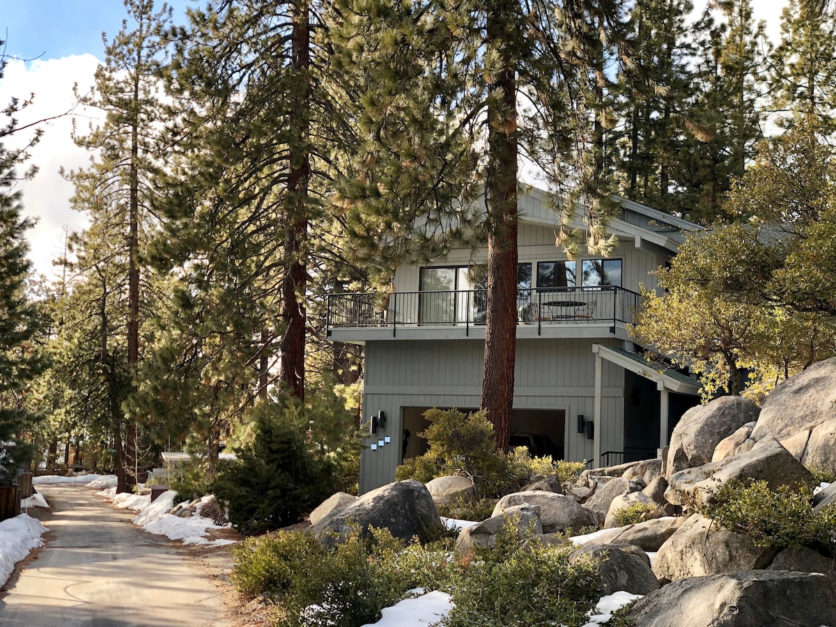 A two-level home is nestled among tall pine trees, showcasing a balance of natural beauty and modern architecture. The exterior features a deck with glass railings and large windows. Granite boulders and shrubbery are present along the pathway, reflecting a serene mountain setting.