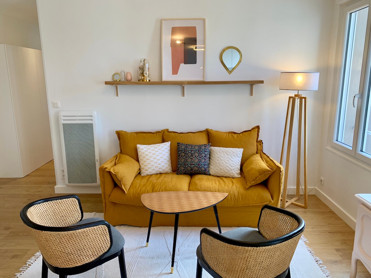 A bright living area features a mustard yellow sofa, adorned with various decorative cushions. Two black and rattan accent chairs are positioned around a circular wooden coffee table. Soft natural light enters through a large window, highlighting the minimalistic decor above the sofa.