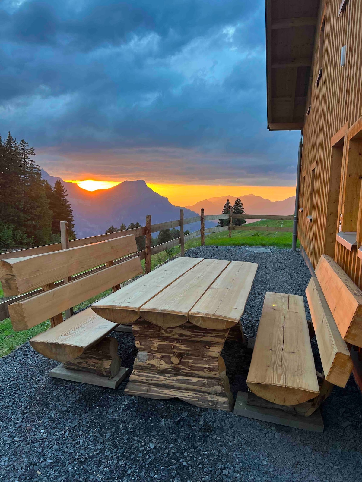 A wooden picnic table and benches are positioned outside the house, offering views of the sunset over the mountains. The table is crafted from natural logs, surrounded by gravel underfoot, and framed by lush trees in the background.