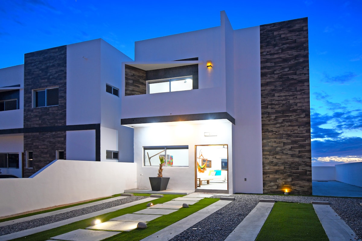 A modern home exterior is presented at dusk, featuring a combination of stone and smooth white walls. A well-maintained pathway, accented by recessed lighting, leads to the entrance. Large windows invite natural light, while a decorative planter adds a touch of greenery.