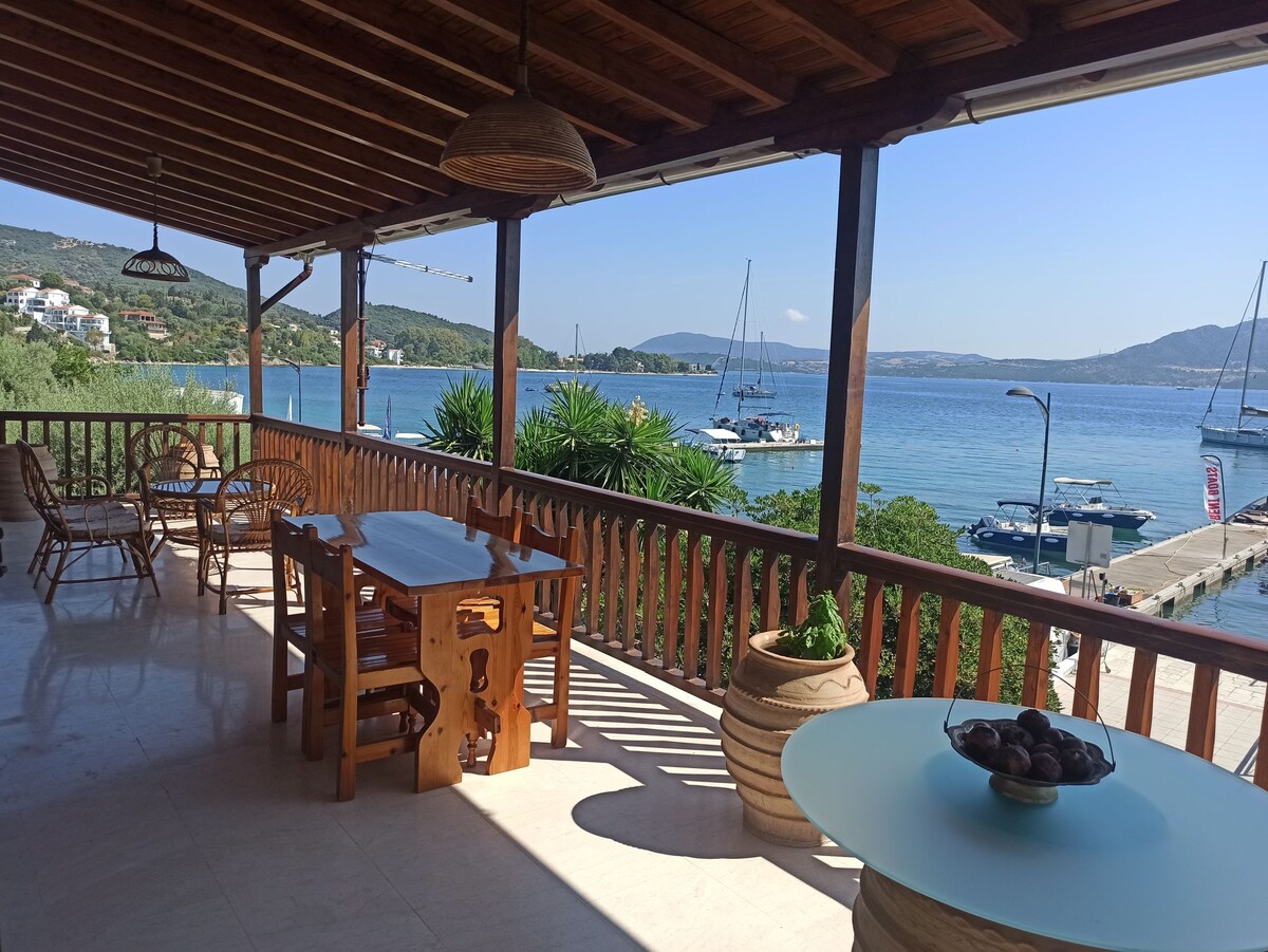 A spacious balcony offers a dining table and seating area, with wooden furnishings enhancing the natural atmosphere. Scenic views of the sea are visible, featuring boats in the distance and lush greenery. The space is well-lit, creating an inviting environment for relaxation.