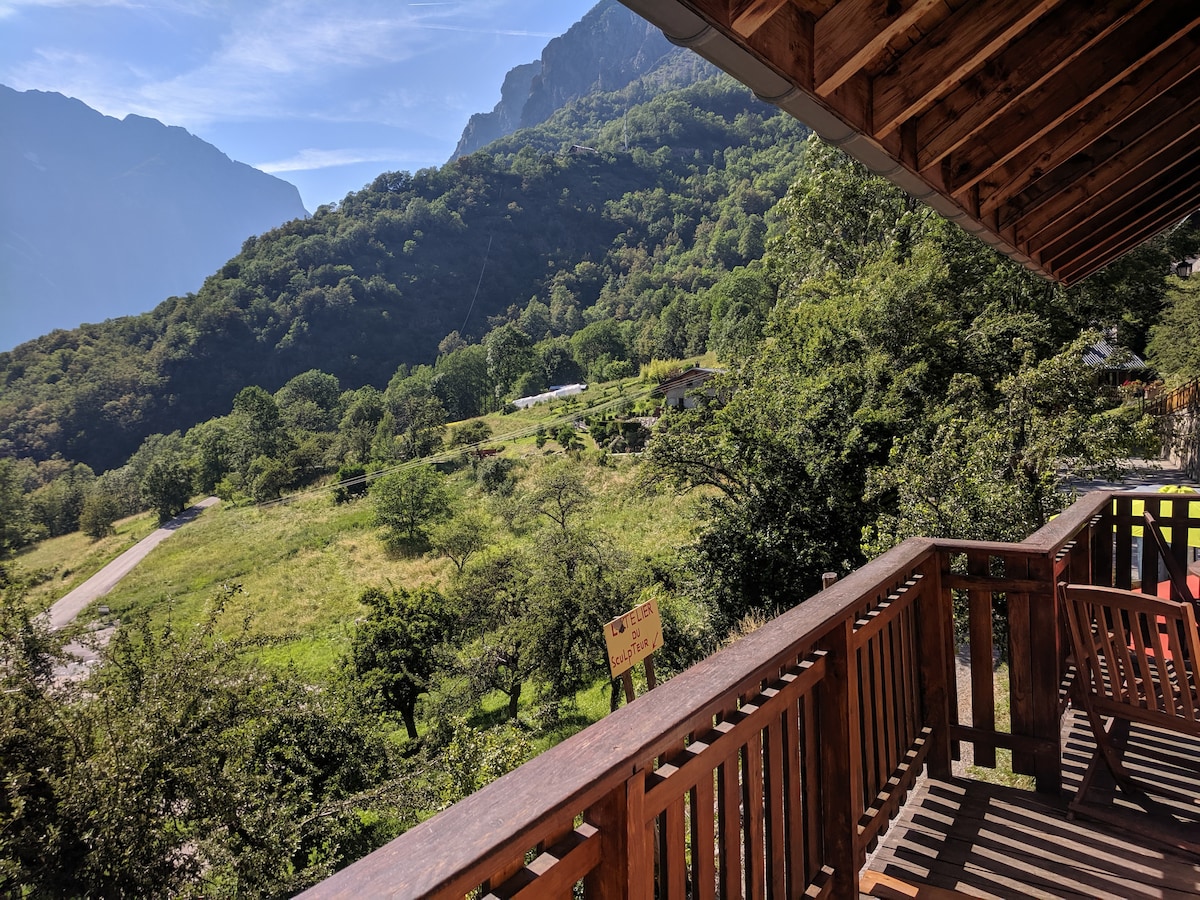 A wooden balcony overlooks a lush green landscape, dotted with trees and meadows. The backdrop features rolling hills and mountains under a clear blue sky, creating a serene outdoor environment. Sunlight bathes the area, enhancing the view of nature.