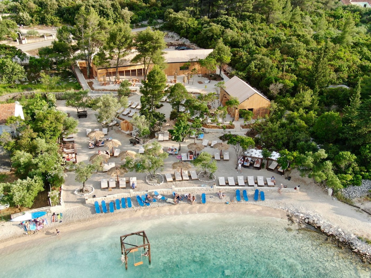An aerial view captures a beach scene lined with sunbeds and umbrellas beside calm, clear waters. A wooden structure sits amidst greenery, and pathways weave through the landscape. A sandy beach is dotted with guests enjoying the serene environment.