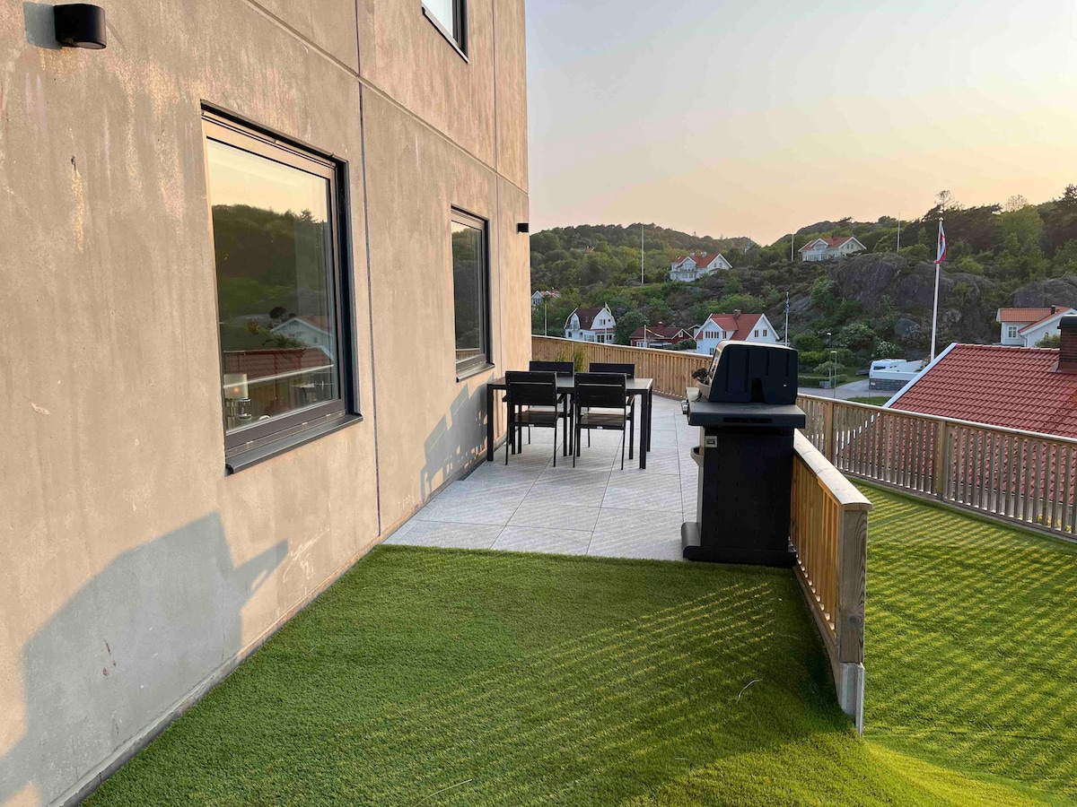 An outdoor terrace features a dining table with six chairs, accompanied by a barbecue grill. The area is surrounded by lush green grass, with views of nearby trees and rooftops in the distance, illuminated by the warm glow of sunset.