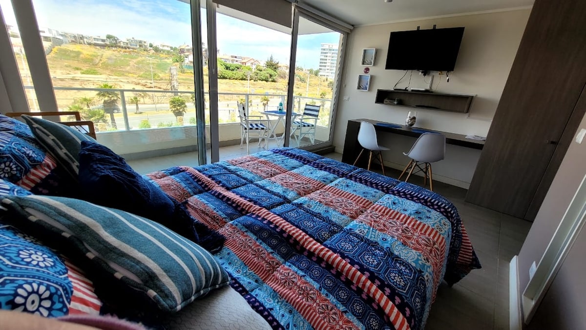 A spacious bedroom features a vibrant patterned blanket laid over a large bed. A desk and chairs are positioned nearby, with a wall-mounted television above. Large windows offer unobstructed views and natural light, and a private terrace is visible with outdoor seating.