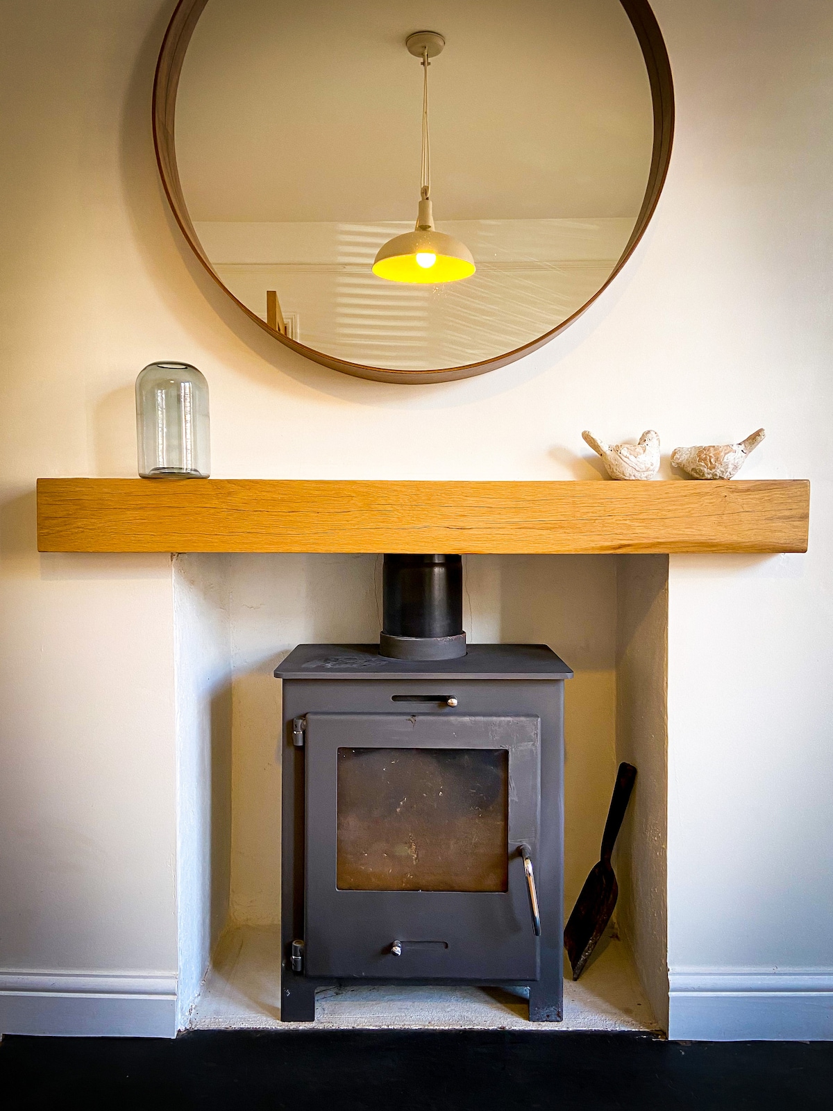 A modern wood burner is positioned within a minimalist alcove, accentuated by a wooden mantel above. A round mirror reflects soft light from a pendant fixture. Two decorative birds and a glass jar adorn the mantel, enhancing the simple aesthetic.