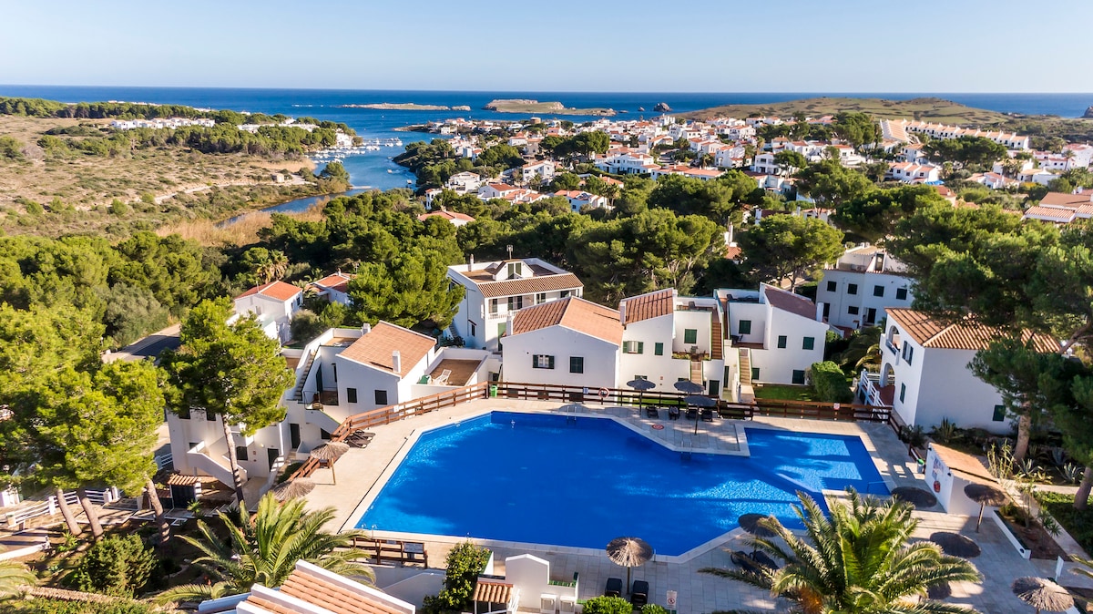 A spacious community pool is surrounded by palm trees and landscaped gardens. The clear blue water reflects sunlight, while nearby buildings feature white facades with red-tiled roofs. A serene view of the coastline and greenery can be seen in the distance, enhancing the tranquil environment.