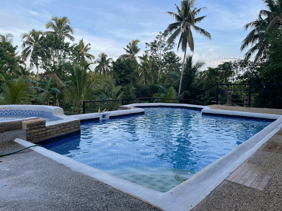 A serene swimming pool is framed by lush tropical greenery, with palm trees visible in the background. The pool features a shallow area and a smooth surface, reflecting the sky above. The patio area is bordered by a rustic railing, enhancing the peaceful surroundings.
