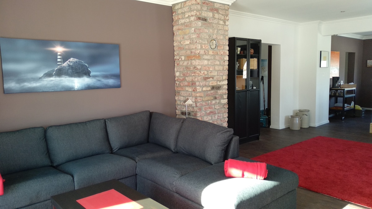 A spacious living room features a large sectional sofa in a dark tone, complemented by vibrant red accents. A stone wall adds texture, while a large framed photograph of a lighthouse hangs above. Natural light floods the space, highlighting the warm atmosphere.