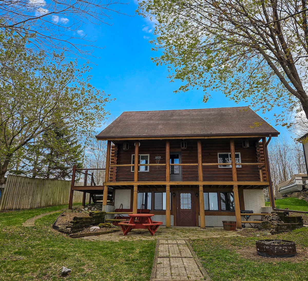 A spacious log cabin is presented, featuring a two-story structure with a wooden deck extending from the upper level. A stone pathway leads to a patio area with a picnic table and firepit, surrounded by grassy lawn and mature trees under a clear blue sky.