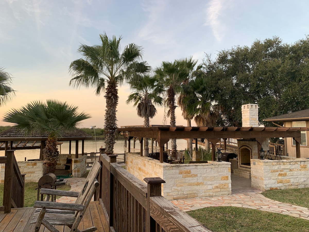 An outdoor space is framed by tall palm trees, showcasing a covered patio area with stone walls and a built-in fireplace. In the background, the water is visible, reflecting the warm hues of sunset, while wooden seating provides areas for relaxation.