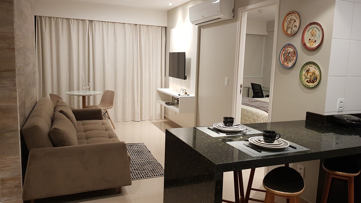 A contemporary living space is presented, featuring a comfortable sofa paired with a stylish black dining table set for two. A flat-screen TV is mounted above a white media console. Decorative plates adorn the wall, while large curtains cover the floor-to-ceiling window, allowing soft light to filter in.