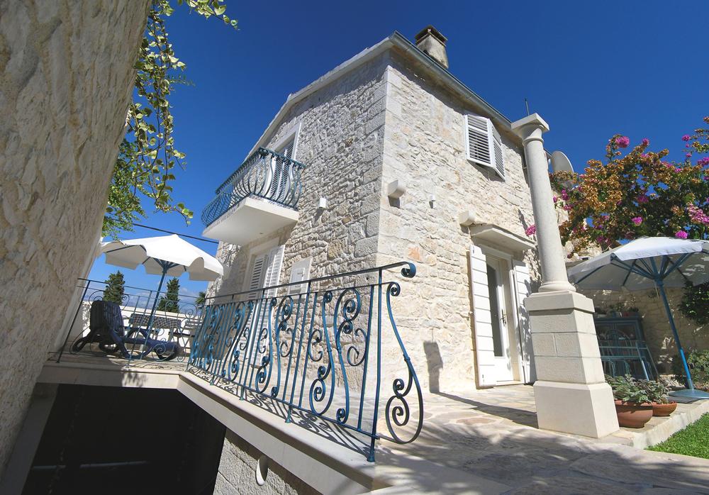 The exterior of the stone house showcases traditional architecture, featuring a wrought iron balcony with blue railings on the upper floor. The surrounding area includes lush greenery and outdoor seating shaded by umbrellas, contributing to a relaxing outdoor space.