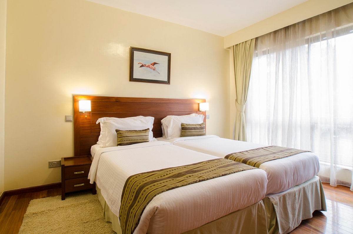 A well-appointed guest room features two spacious beds with neutral-colored linens. A decorative piece hangs above the headboard, while soft natural light filters through sheer curtains. A wooden nightstand is positioned beside each bed, adding functionality to the inviting space.