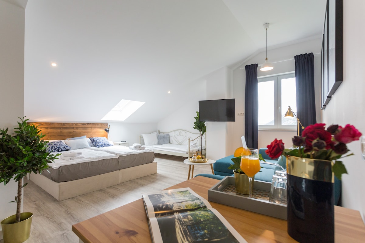 A bright and airy room features a spacious bed with wooden accents, complemented by a cozy seating area. A large window allows natural light to enter, illuminating a small table with a tray of drinks and flowers, adding a touch of elegance to the space.