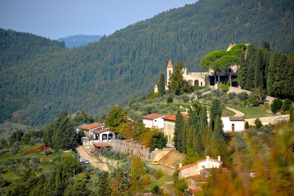 A scenic view captures the estate nestled in the hillside, featuring a blend of stone buildings and lush greenery. The picturesque landscape includes olive trees and paved paths, with a historic watchtower visible in the distance, reflecting the region's rich heritage.