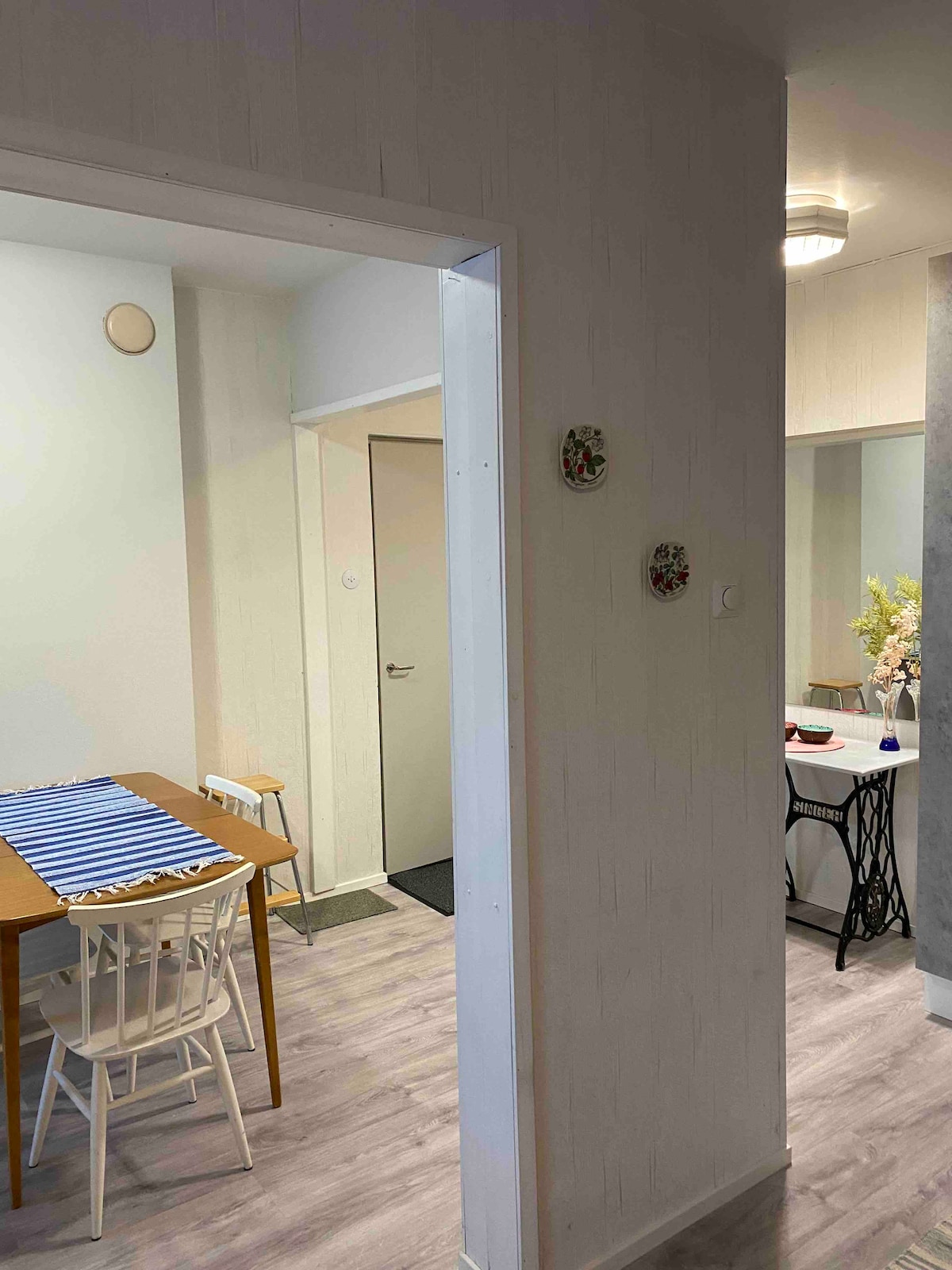 A light-filled corridor connects two inviting dining areas. One side features a wooden table with a blue-striped runner and white chairs, while the other showcases a smoothly styled table with decorative items. The walls are adorned with floral accents, enhancing the welcoming environment.