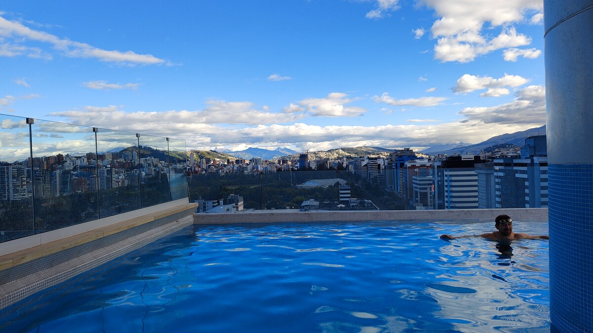 A rooftop pool is featured, surrounded by a transparent barrier offering unobstructed views of the city skyline and distant mountains. The clear water reflects the blue sky dotted with clouds, creating a serene ambiance for relaxation.
