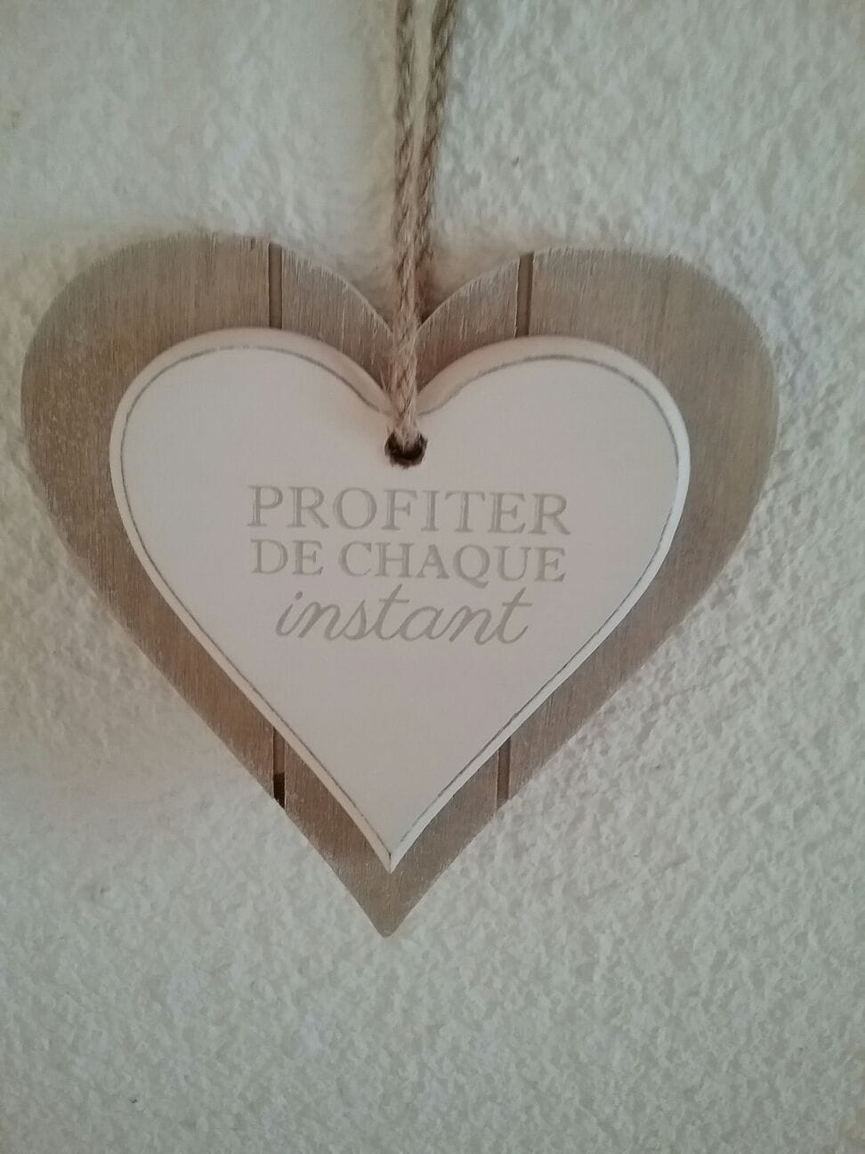 A wooden heart-shaped sign hangs on a textured wall. The sign features the phrase 'PROFITER DE CHAQUE instant' imprinted in a clear font, providing a warm decorative touch to the interior.