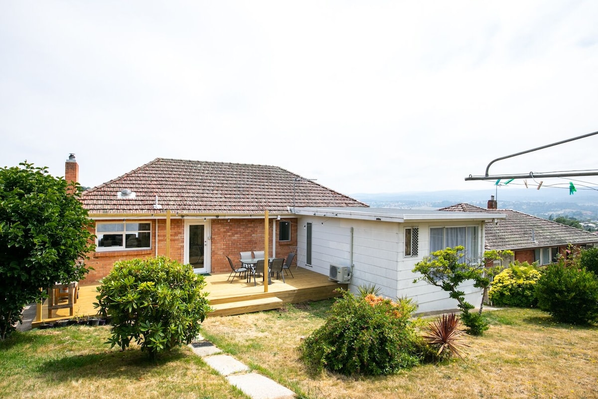 A spacious home with a large deck is visible, offering an outdoor dining area. Lush greenery surrounds the property, and the facade features a combination of brick and wood. Scenic views of the landscape can be seen in the background.