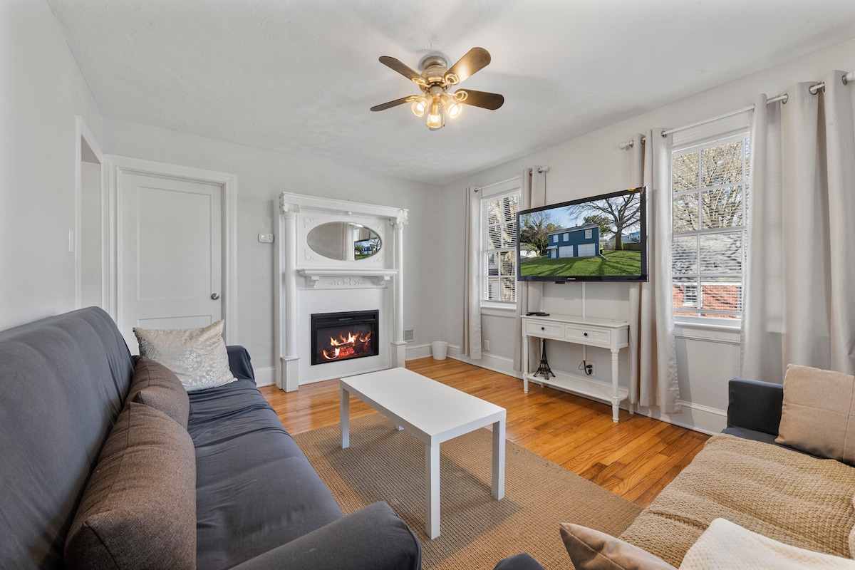 A cozy living area features a plush couch, warm wood flooring, and a modern electric fireplace. A large TV is mounted on the wall, displaying an exterior view of the cottage. Natural light fills the room through multiple windows, complemented by soft curtains.
