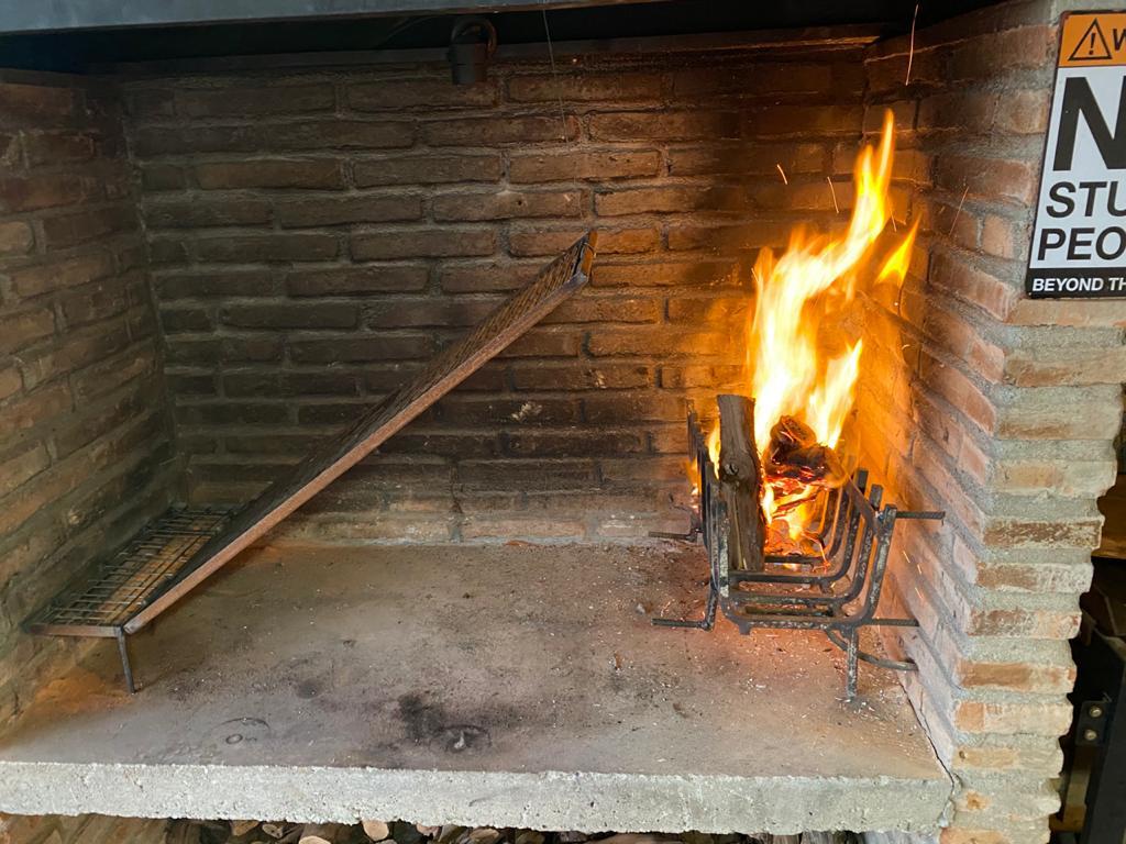 A traditional outdoor grill, featuring a stone and brick structure. Flames are visible, rising from the firewood placed on the grill. A metal grate is positioned to the left, allowing for cooking or heating. The background showcases a warm, rustic ambiance.
