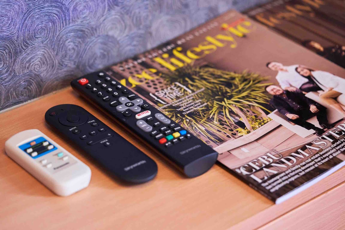 An assortment of remote controls is placed neatly beside a lifestyle magazine on a wooden surface. The magazine's cover features a vibrant photograph of a group enjoying leisure time in a garden setting, surrounded by lush greenery.