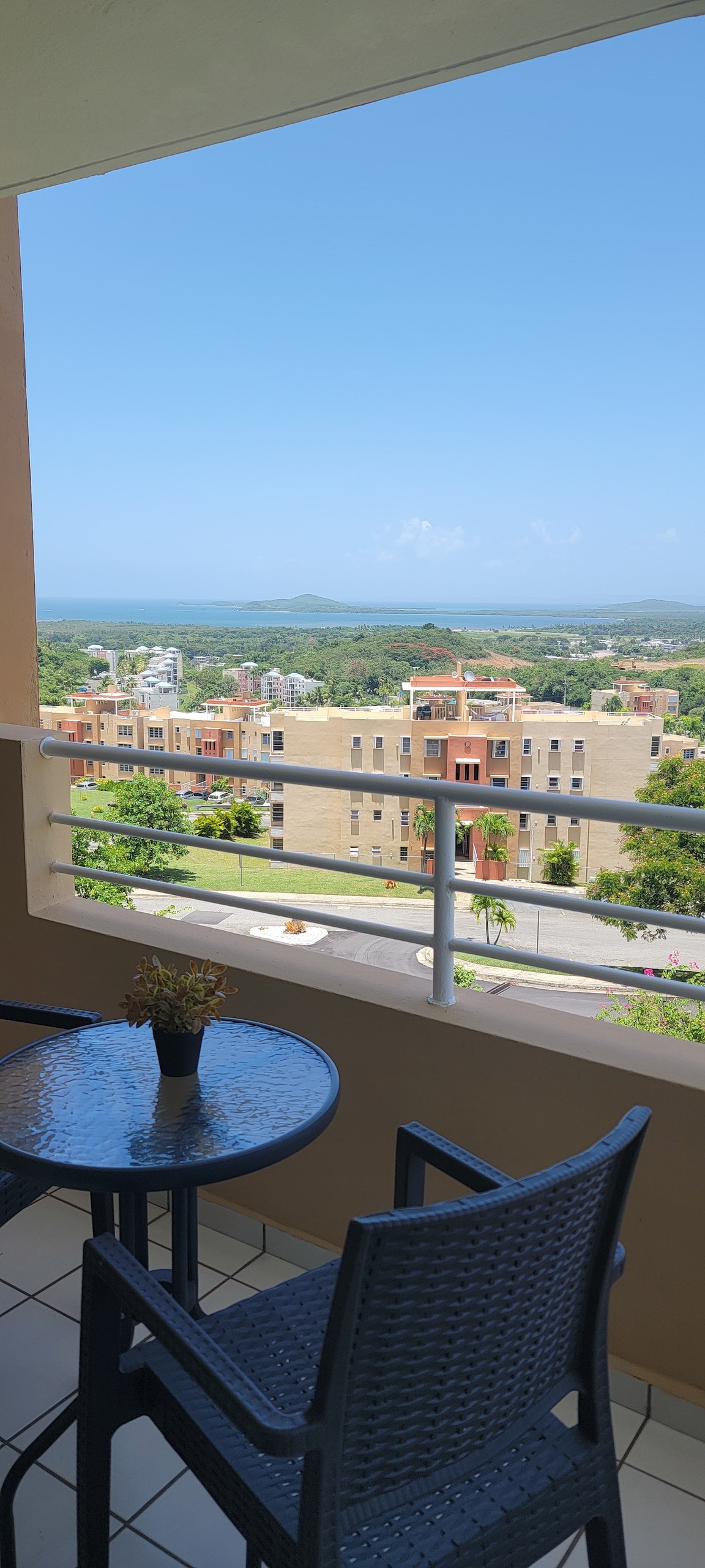 A balcony view features a glass-topped table with two wicker chairs, overlooking a serene landscape that includes buildings and lush greenery. The clear blue sky enhances the peaceful ambiance, inviting relaxation in the outdoors.