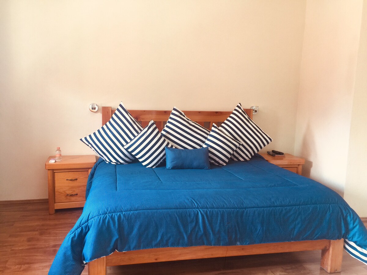 A spacious bed is centered against a light-colored wall, adorned with striped pillows and a solid blue accent pillow. Two wooden nightstands flank the bed, offering a warm contrast to the blue bedding. Natural light enhances the inviting ambiance.
