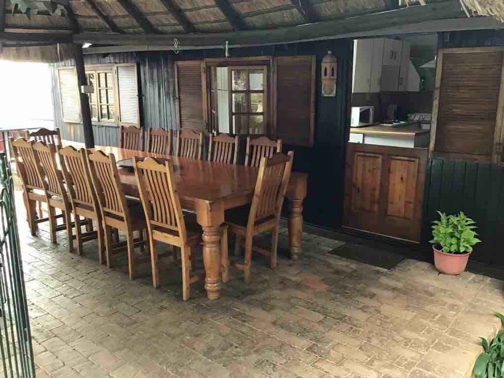 A spacious dining area features a large wooden table surrounded by multiple chairs. The rustic interior design includes thatched roofing and wooden accents, providing a cozy environment. A doorway leads to a kitchen area, enhancing the inviting communal space.