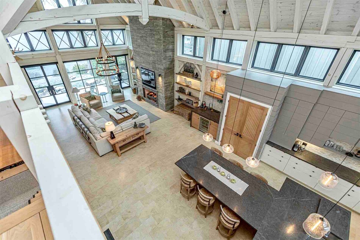 An aerial view captures a spacious open-concept living area with a combination of modern and rustic design elements. Large windows allow natural light to flood the space, which includes a cozy seating area, a dining table with four chairs, and a well-appointed kitchen featuring a large island and pendant lighting.