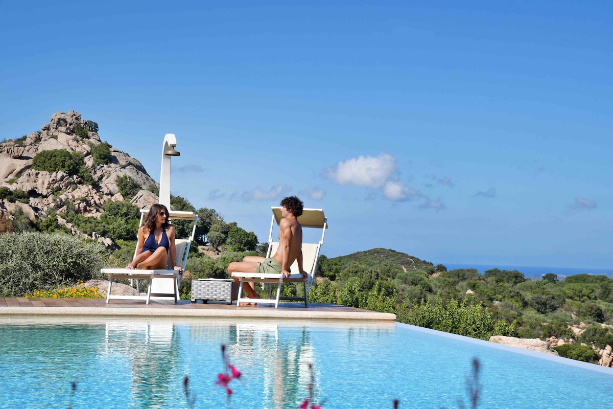 Sun loungers are positioned beside a tranquil infinity pool, offering views of the surrounding hills and distant sea. The clear blue sky enhances the serene atmosphere, with lush greenery framing the scene. Two individuals relax, enjoying the peaceful setting.