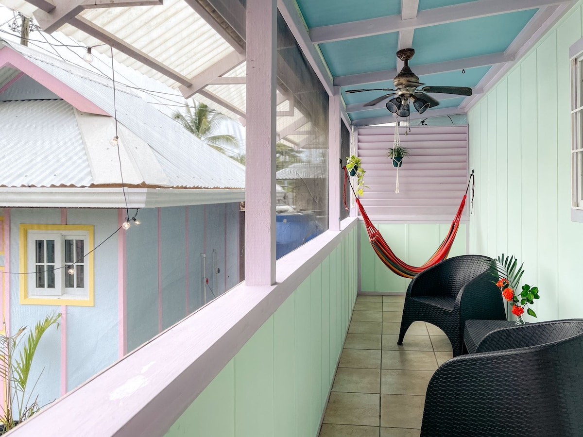 A charming outdoor balcony features comfortable seating and a vibrant hammock, surrounded by colorful walls in soft pastel hues. A ceiling fan provides ventilation while natural light filters through nearby windows. The space is designed for relaxation in a tranquil tropical setting.