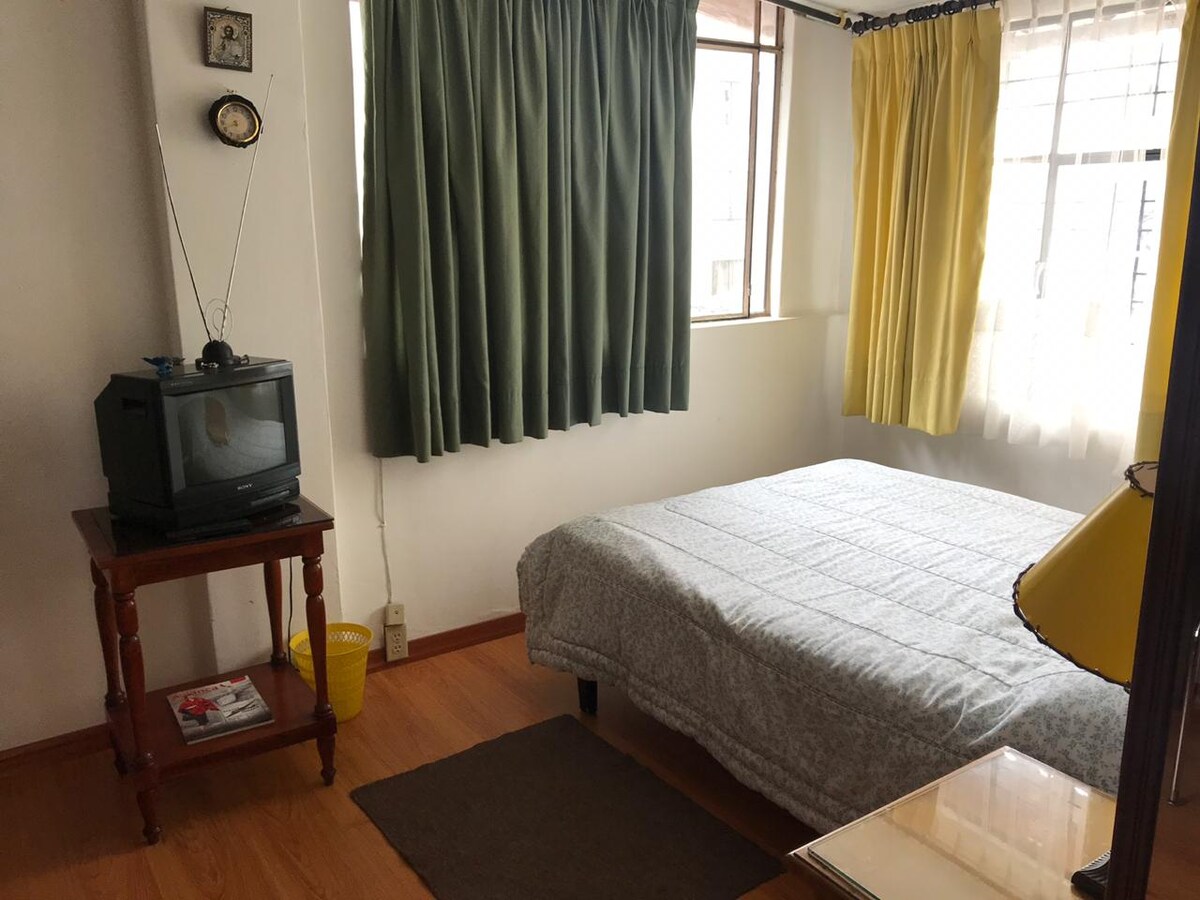 A well-lit sleeping area features a spacious bed covered with a light-colored quilt. Two windows are adorned with yellow and green curtains, allowing natural light to filter in. A small wooden side table and a vintage television are situated beside the bed.