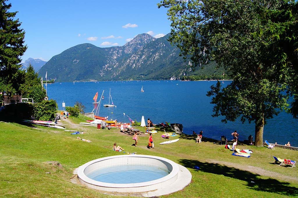 A scenic view of the lake d'Idro features a grassy area leading to the water's edge. Sunbathers are scattered on the grass, with boats visible on the lake. The backdrop showcases mountains under a clear blue sky, adding to the tranquil atmosphere.