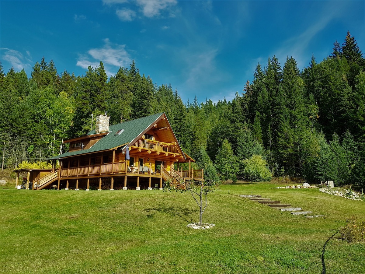 The blockhouse is surrounded by lush green forests and rolling landscapes, featuring a large deck that spans the front. Steps lead down through the yard towards the natural terrain, with the peaceful backdrop of trees and blue sky providing a serene atmosphere.