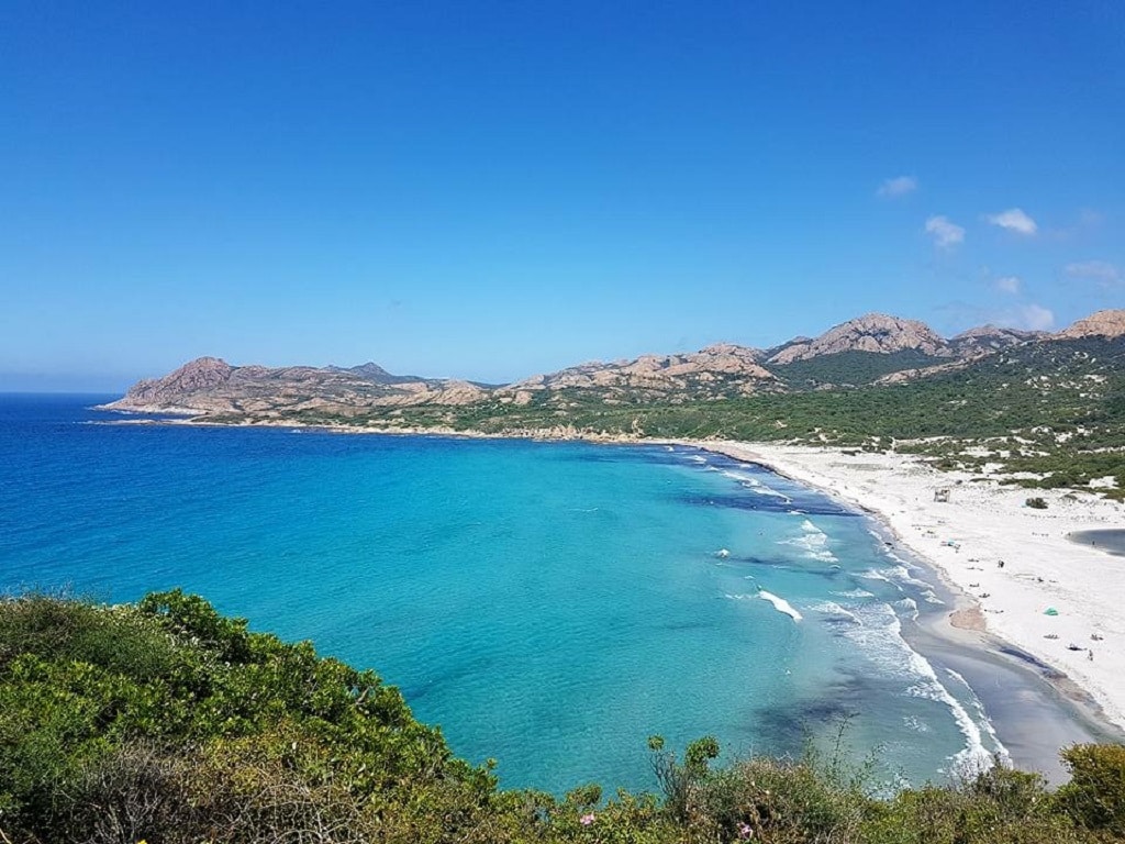 A sweeping coastal view captures the vibrant turquoise waters meeting a sandy shore, bordered by rugged mountainous terrain. The bright blue sky enhances the serene landscape, inviting relaxation and exploration along the coastline.