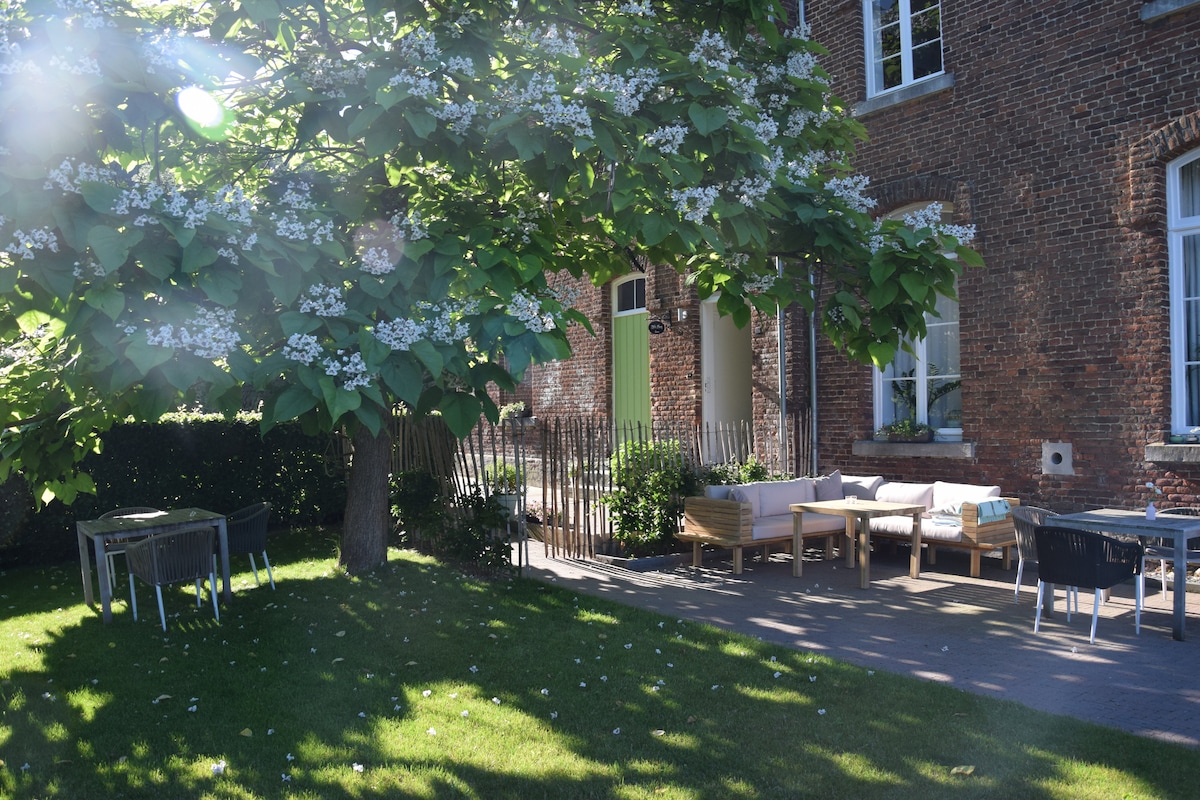 A spacious outdoor area features a well-maintained lawn and shaded seating under a large tree. Various seating arrangements, including wooden benches and tables, provide options for relaxation. The brick exterior of the building adds to the inviting atmosphere of the space.