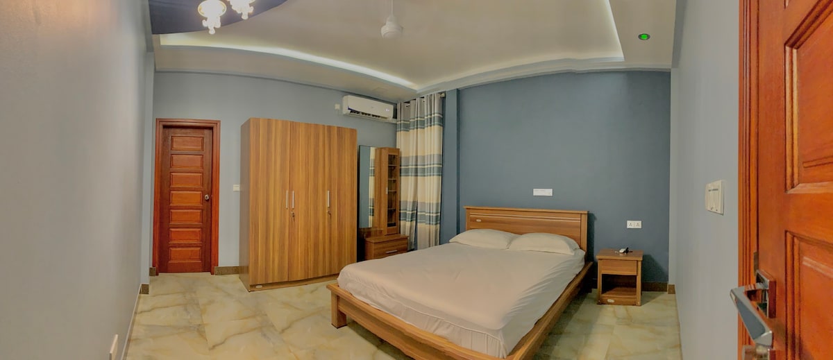 A spacious room features a comfortable bed with neutral bedding, complemented by wood furniture. A large wardrobe and bedside table are included. Natural light enters through a window dressed with light curtains, while the walls are painted in a calming blue hue.