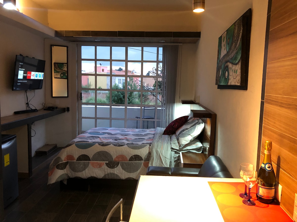 A cozy room features a double bed with a patterned quilt, complemented by a small dining table and two chairs. A wall-mounted TV is positioned near a large window, allowing natural light to fill the space. An artwork hangs above the bed, adding a touch of color.