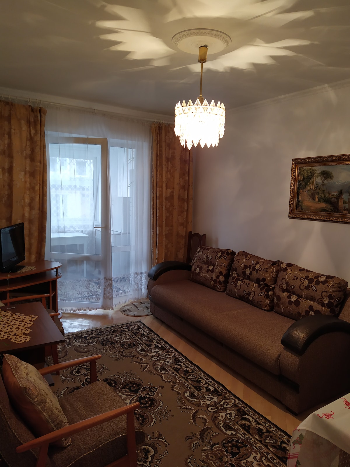 A cozy living area is furnished with a patterned sofa and an armchair, complemented by a wooden coffee table. Natural light filters through sheer curtains, illuminating the space. A decorative chandelier hangs from the ceiling, and a small television is positioned on a corner unit.