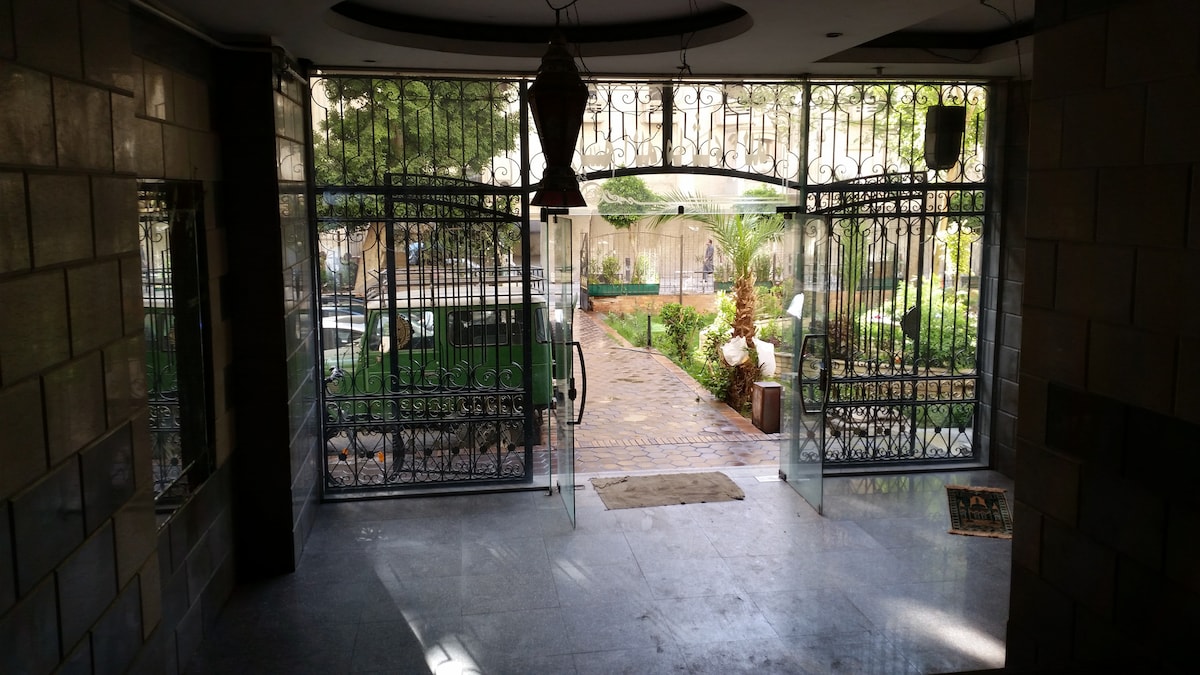 An ornate gated entrance is seen, leading to a small garden area. The floor is tiled and features a slight sheen. Through the gates, greenery is visible with well-maintained pathways that guide through the outdoor space.