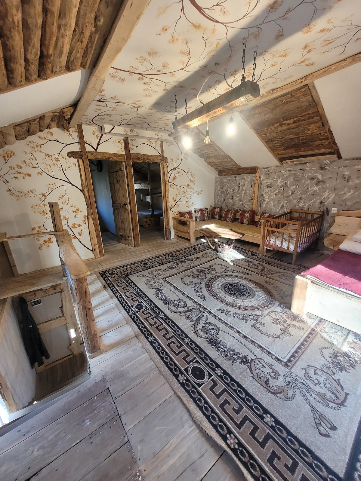 The interior features wooden beams and a decorative ceiling, complemented by floral-patterned wallpaper. A patterned area rug covers the wooden floor, while a spacious bed and a set of other seating options provide comfort. Light fixtures hang from the ceiling, enhancing the warm atmosphere.
