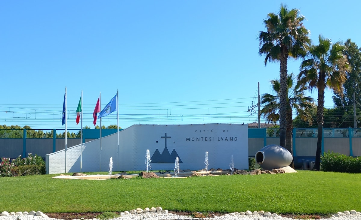 The welcoming entrance of Montesilvano is showcased, featuring a large sign with the city's name and symbolic imagery. A manicured lawn surrounds the sign, complemented by fountains and palm trees. Flags representing various regions are displayed prominently, indicating a vibrant community.