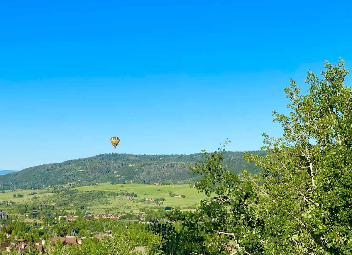 A vibrant hot air balloon is seen floating above rolling green hills, framed by lush quaking aspens. The clear blue sky enhances the scenic view, highlighting the beauty of the surrounding landscape.